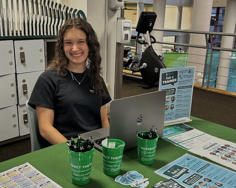 A personal trainer smiles and sits behind the Ask a Trainer table.