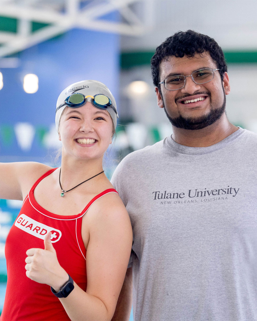 Smiling lifeguard in red suit gives thumbs up with a man wearing glasses at the natatorium.