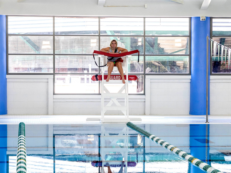 Lifeguard on a high chair at an indoor pool, holding a red rescue tube.