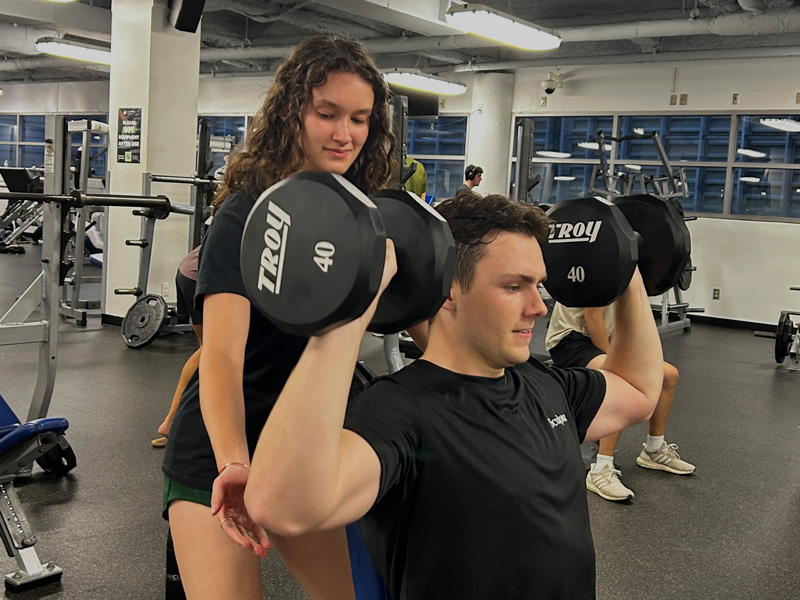 A personal trainer works with a client lifting dumbbells in the weight room.