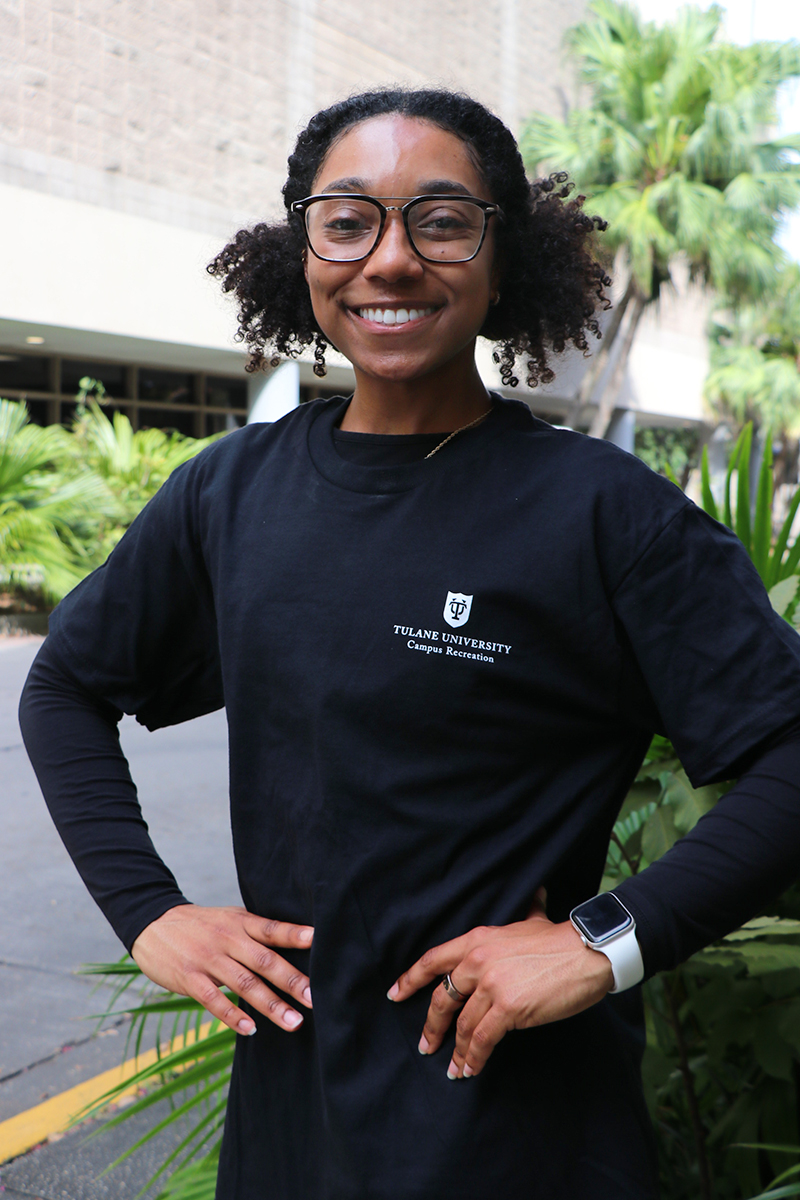 Smiling woman with glasses and curly hair, wearing a black t-shirt with a logo, hands on hips.