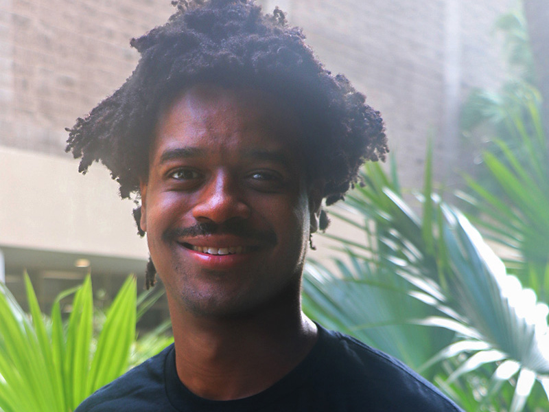 Smiling man with a dark curly afro and mustache, outdoors with plants.