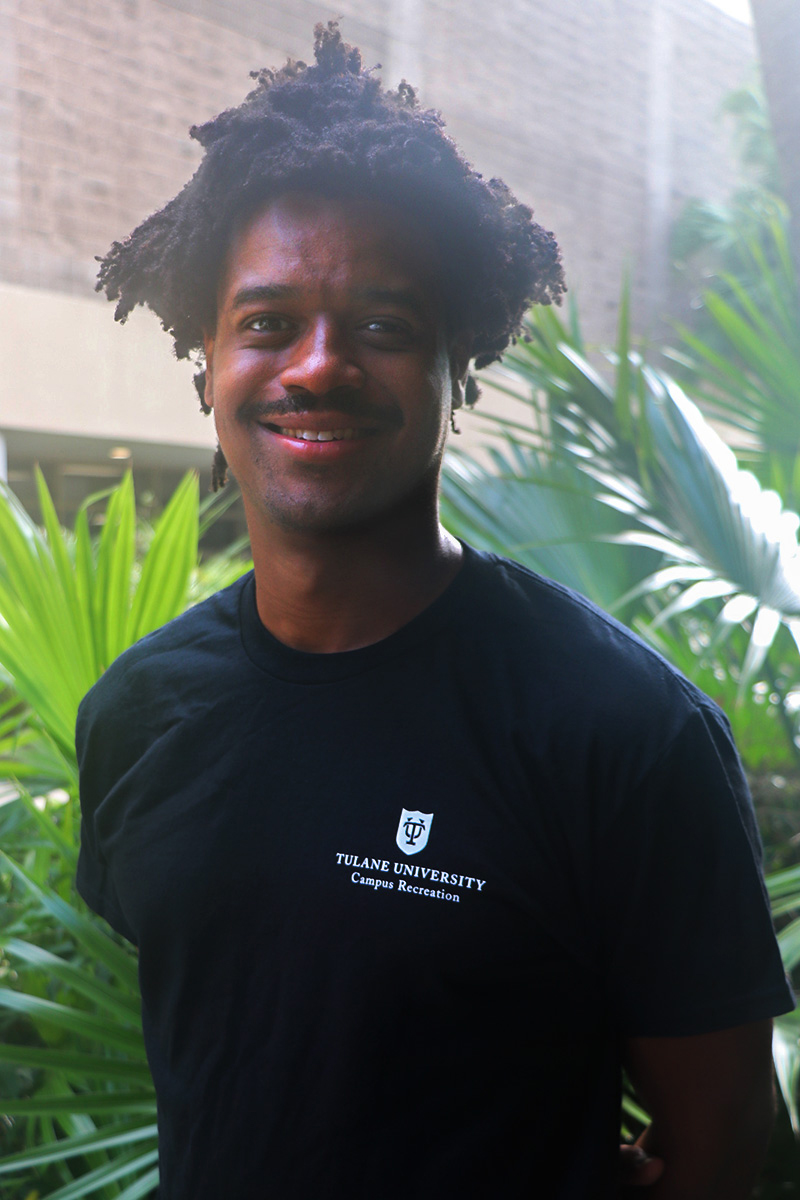 Young Black man with dreadlocks smiling, wearing a dark t-shirt, outdoors with plants.