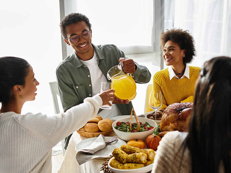 A young man pours a sweet drink from a jug into a friend's cup at a Thanksgiving dinner with bread rolls, salad, corn, and turkey. A young man pours a sweet drink from a jug into a friend's cup at a Thanksgiving dinner with bread rolls, salad, corn, and turkey.